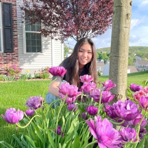 A picture of Wendy in her front yard full of pink flowers and Wendy kneeling on the ground and smiling at the camera.